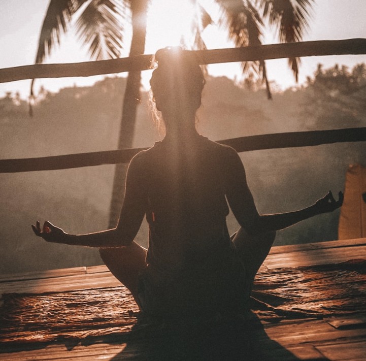 Silhouetted person meditating in lotus pose on a wooden deck at sunrise with palm trees behind